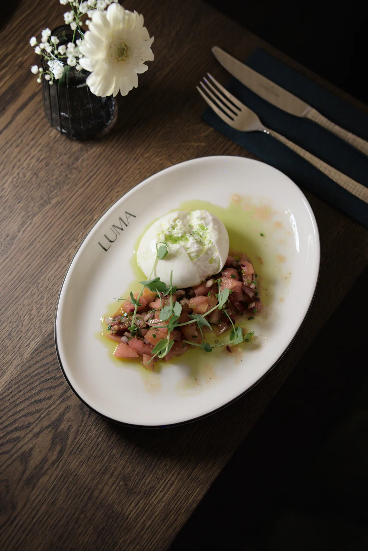 Ein weißer Teller mit Burrata und bunten Zutaten auf einem gedeckten Holztisch